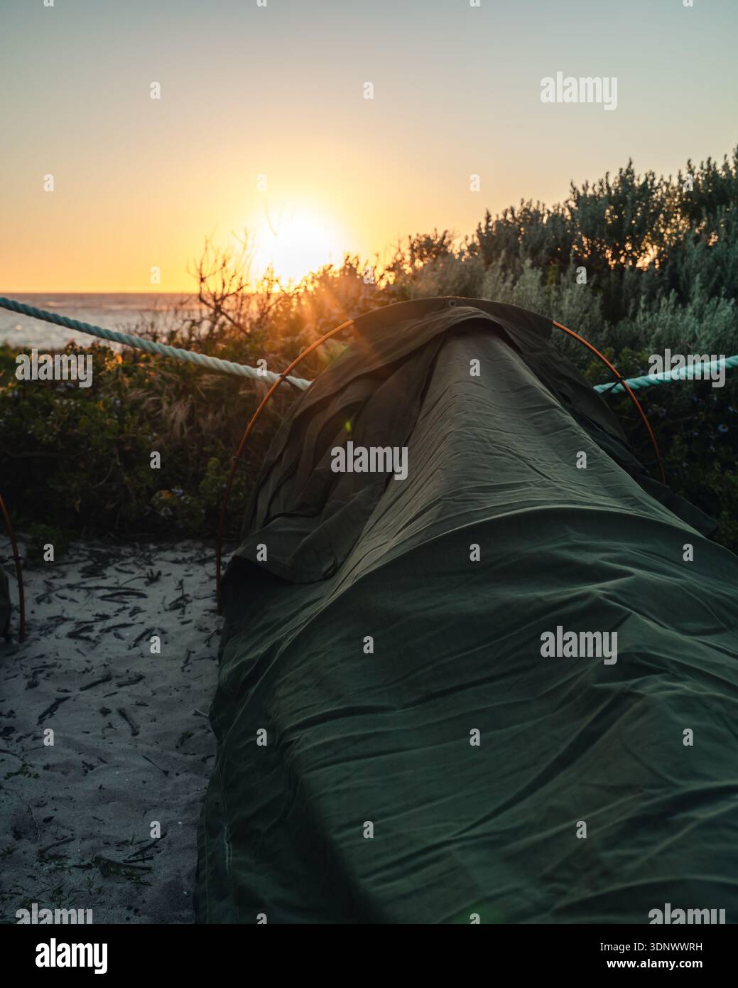 A camping swag rests on sandy ground near coastal vegetation as the sun sets over the ocean. Warm light filters through the shrubs at sunset. Stock Photo