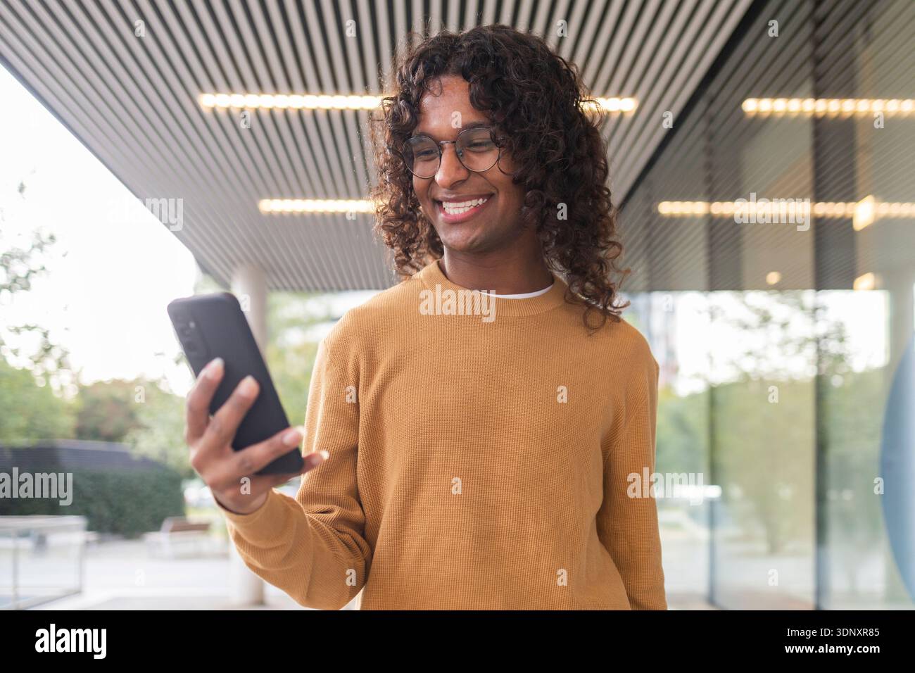 A non-binary individual with curly hair and glasses smiles warmly while using a smartphone under an urban canopy, reflecting a modern and inclusive li Stock Photo