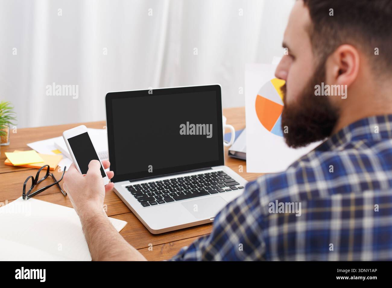 Young beard IT manager looking in mobile phone at work with computer Stock Photo