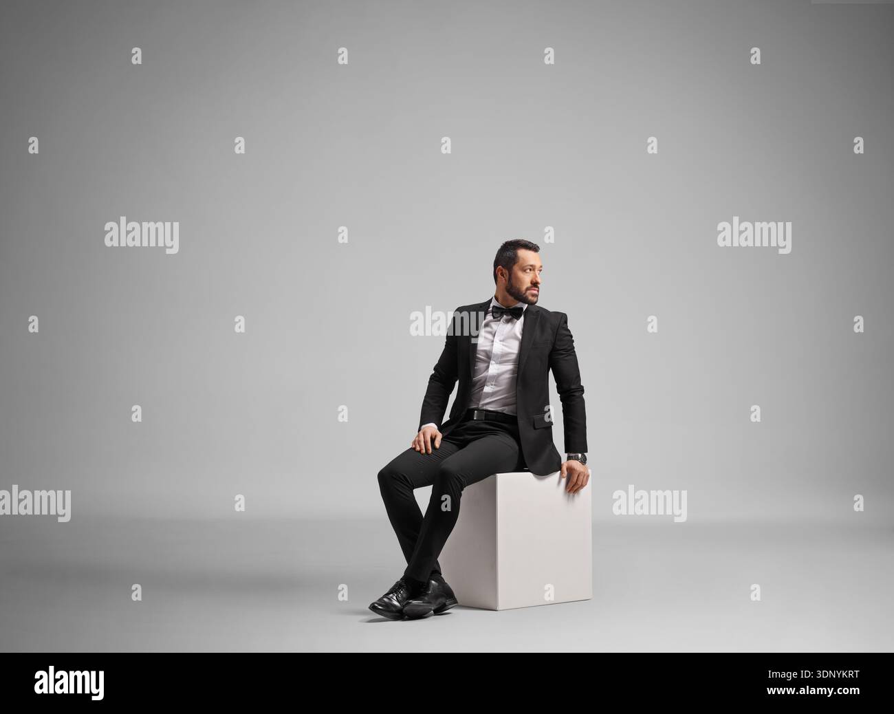 Elegant groom in a black tuxedo sitting on a white cube in a studio Stock Photo