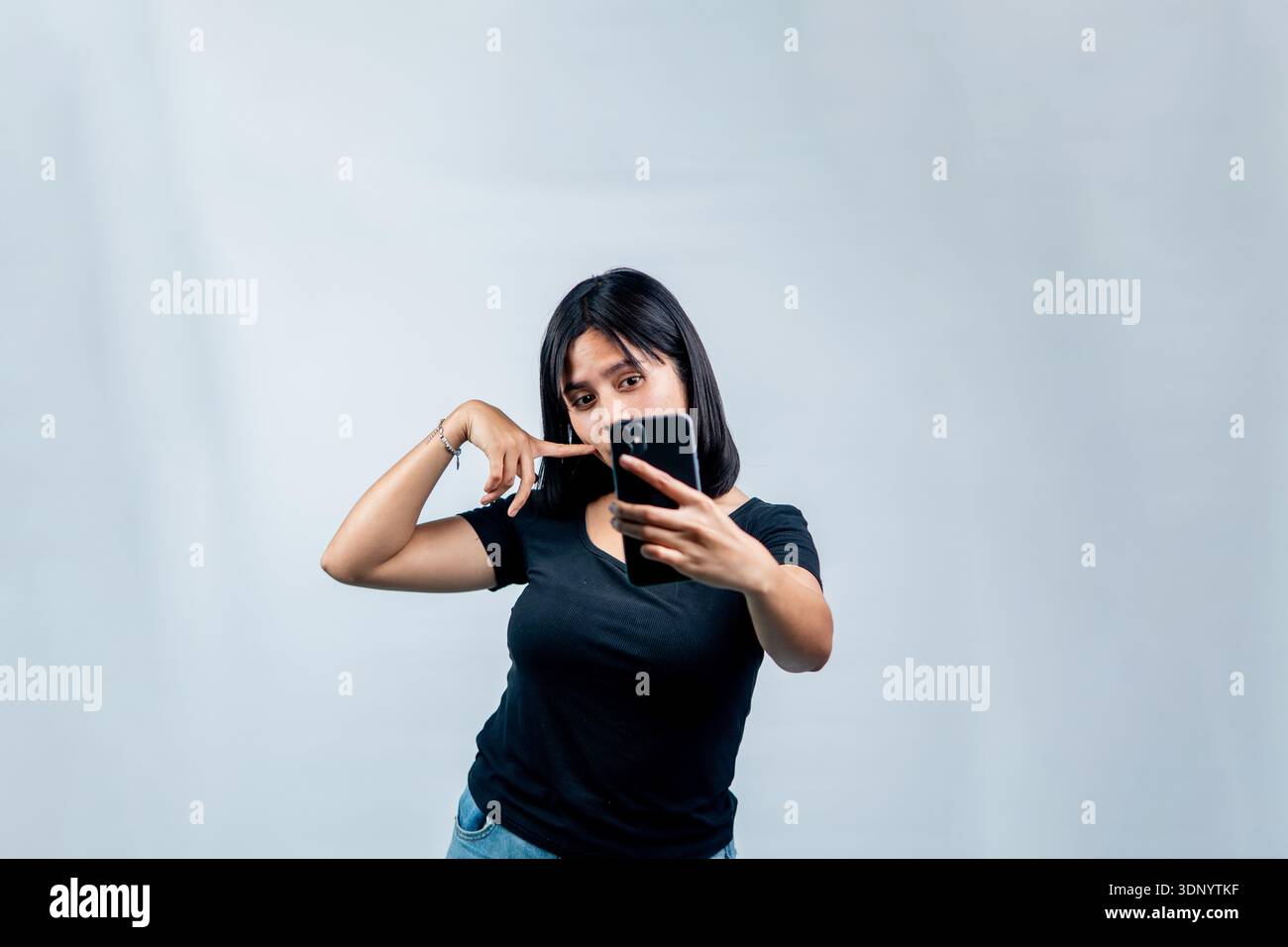 A cheerful young woman in a black t-shirt posing playfully while taking a selfie or recording a video with her smartphone against a white backdrop. Stock Photo