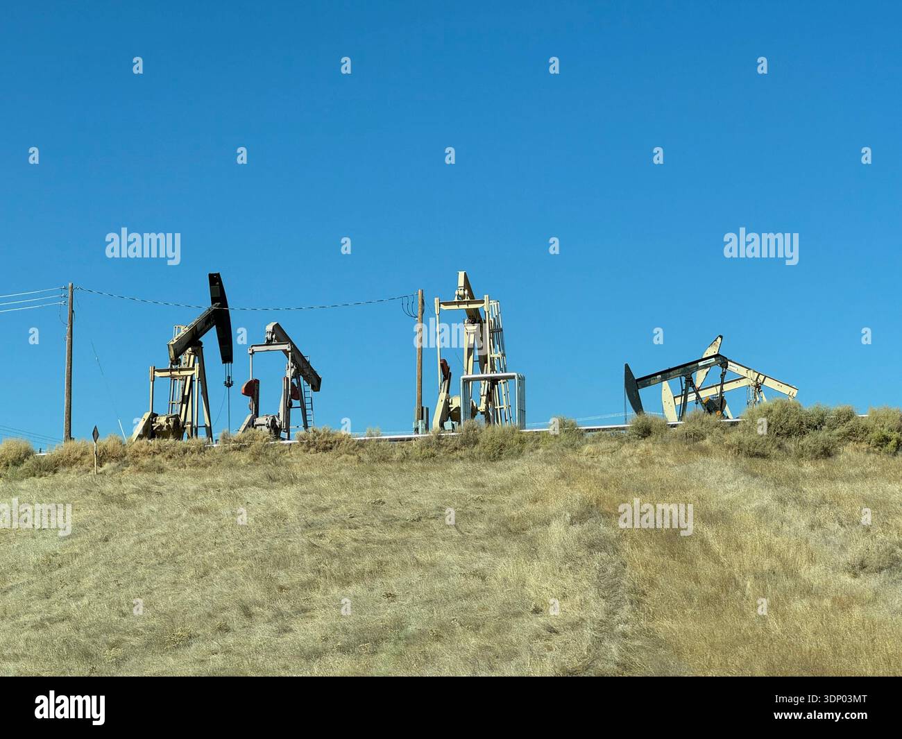 Oil pumpjacks extracting crude oil on a dry grassy hillside under a clear blue sky, petroleum industry machinery in a rural landscape. - Smartphone Captured Stock Image