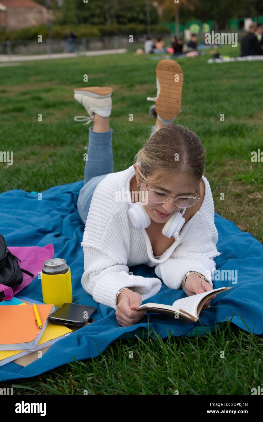Young woman student lying on blue blanket on university lawn, studying outdoors with book, headphones, backpack and drink, relaxed and focused in gree Stock Photo