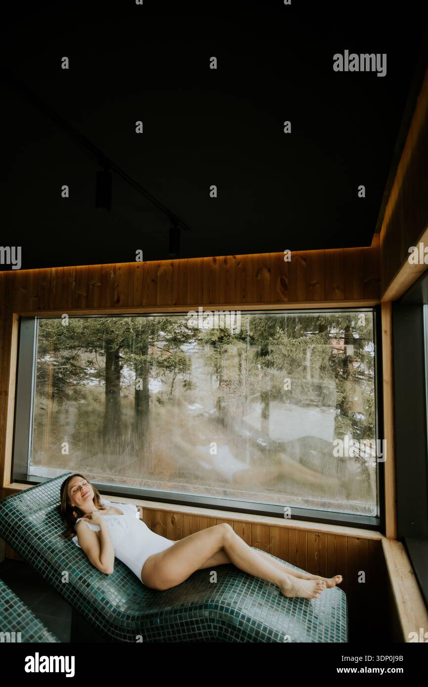 A woman relaxes on a warm stone bed by a big window. She enjoys the ...