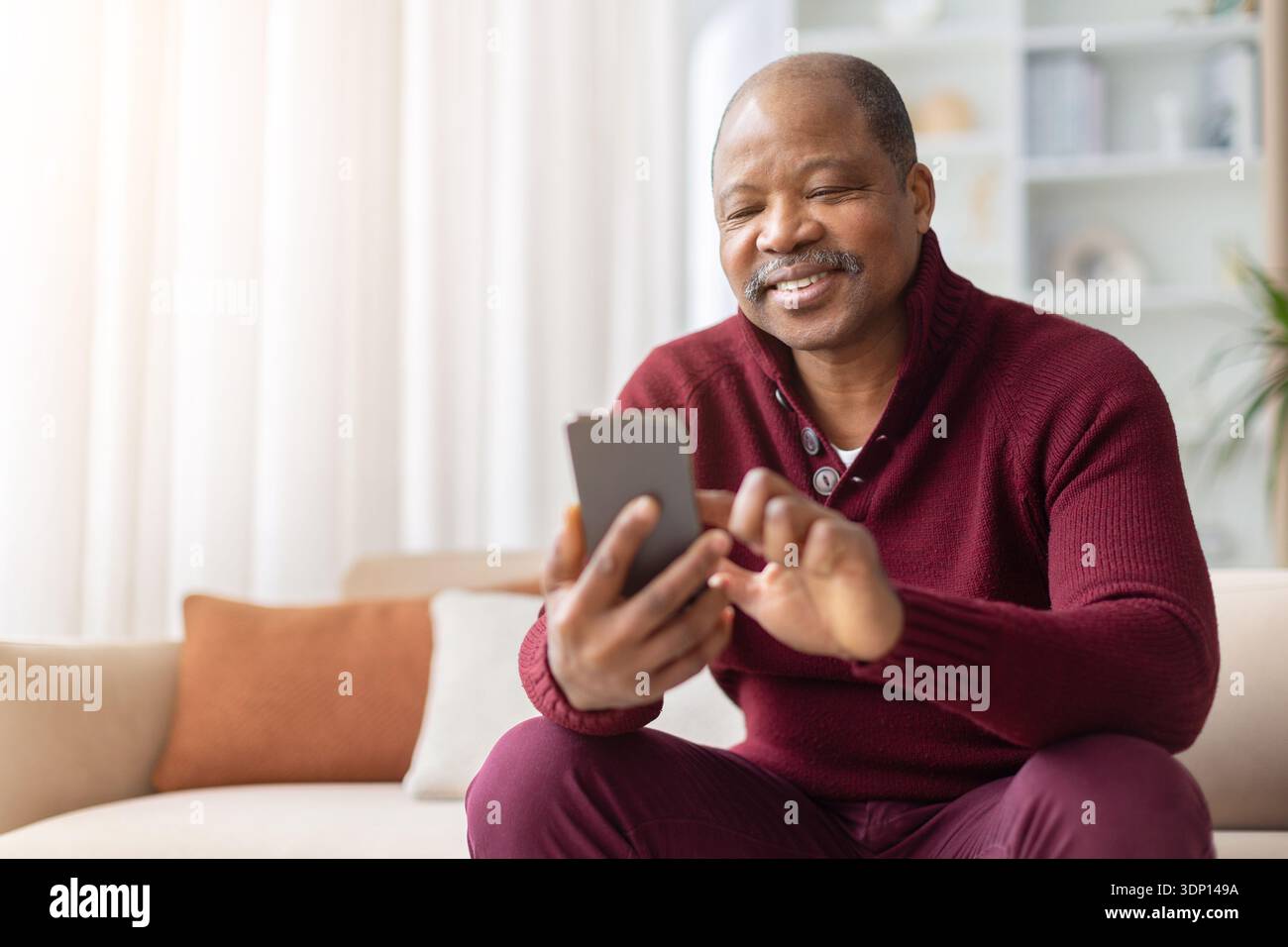Smiling mature black man using smartphone while sitting on sofa at home Stock Photo