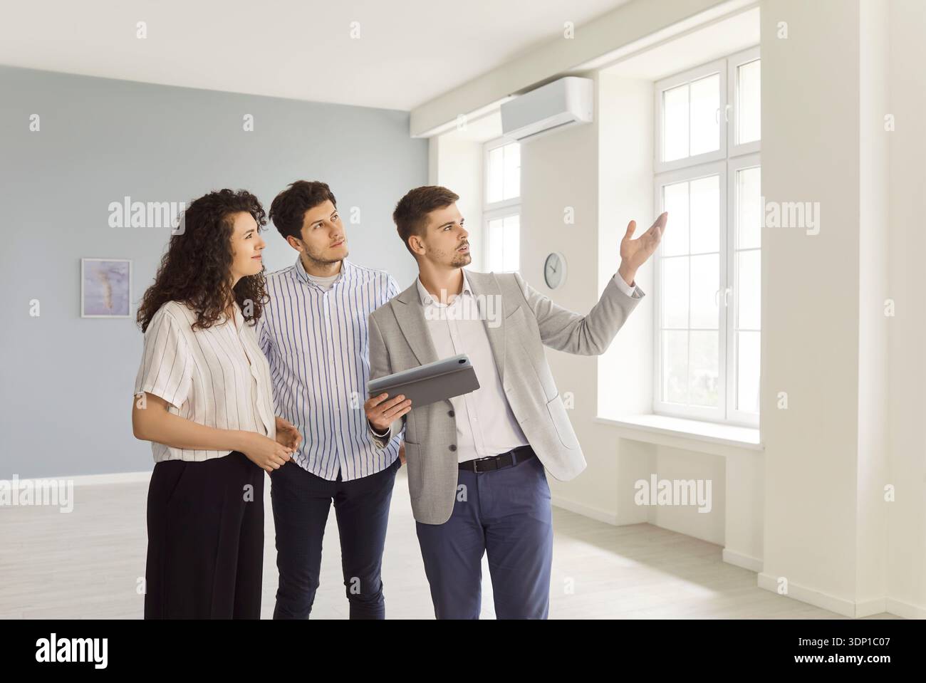 Realtor showing modern house to a young happy couple standing in empty new apartments. Stock Photo