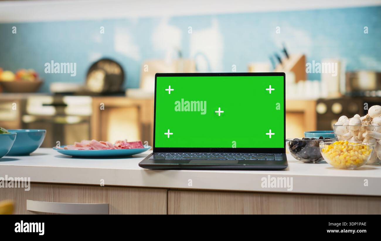 Green screen on laptop in modern home kitchen with cupboards and cookware on display. Countertop holds fresh organic produce and condiments, ideal background for cuisine or healthy eating. Stock Photo