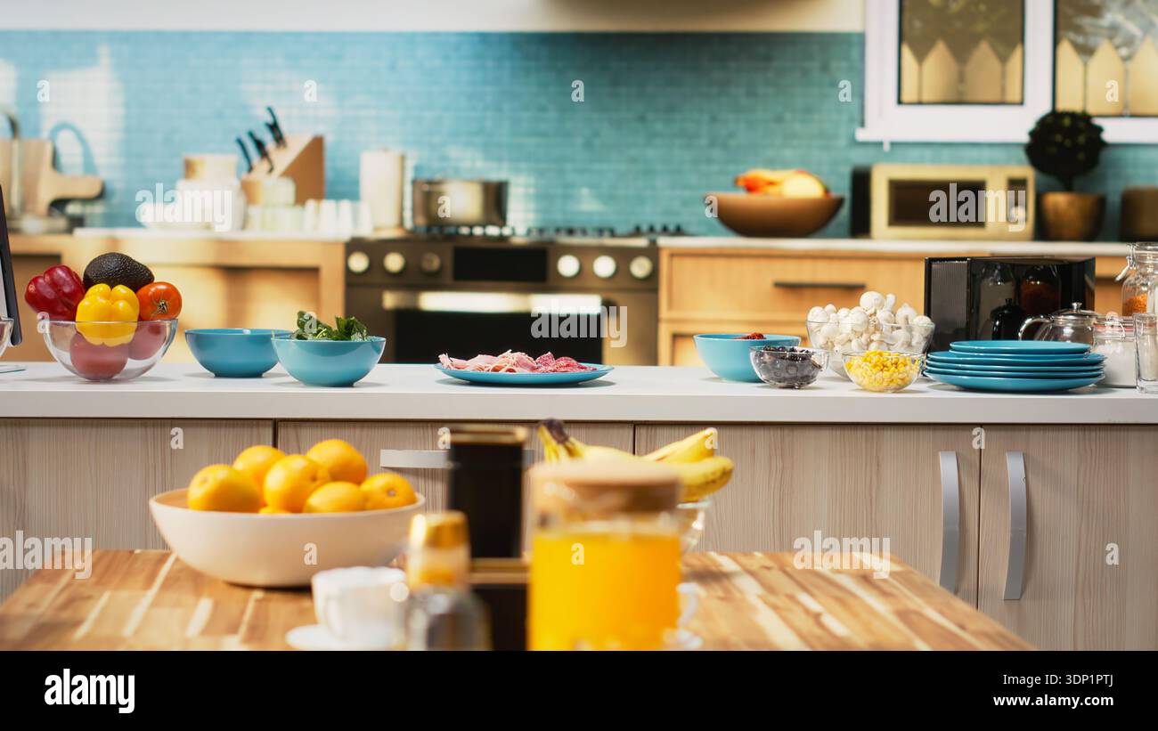 Empty kitchen with wooden dining furniture and modern appliances. Countertop displays colorful vegetables, fresh fruits and spices, representing a stylish home with cozy atmosphere. Stock Photo
