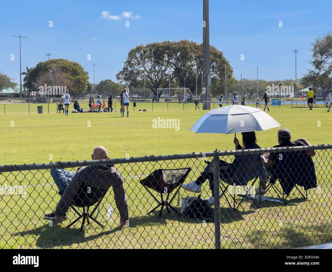 Spectators seated behind a chain-link fence watch a youth soccer match at a sunny outdoor sports field. - Smartphone Captured Stock Image