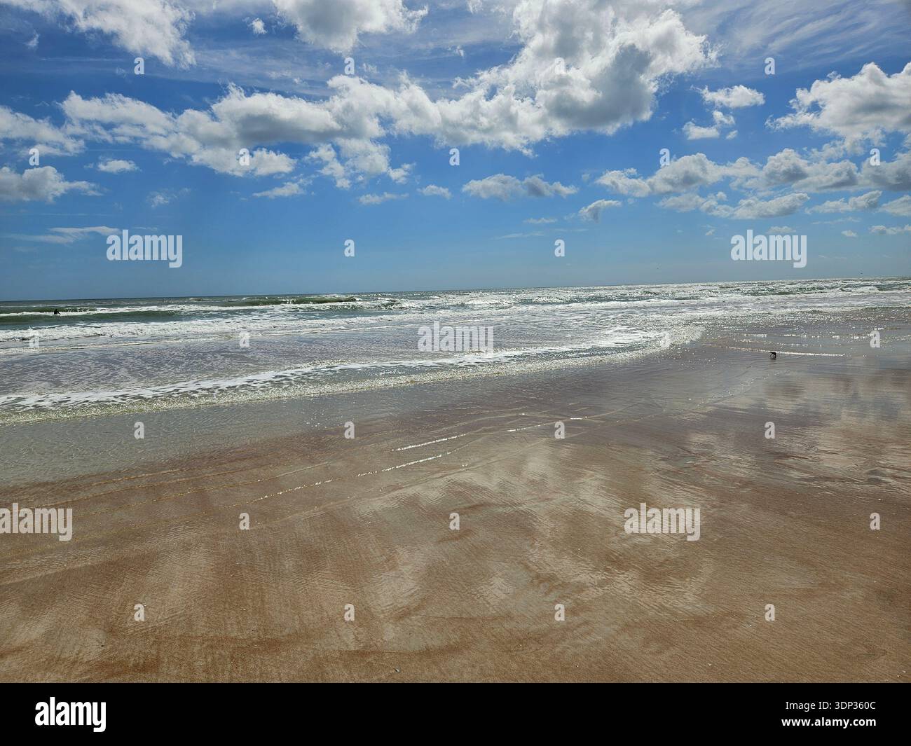 EDITORIAL USE ONLY- DAYTONA BEACH FLORIDA: ​A wide-angle ground-level shot of the Atlantic Ocean coastline in Florida under a bright, cloudy sky. The wet sand creates a mirror-like reflection of the clouds, while gentle white surf rolls onto the shore. - Smartphone Captured Stock Image