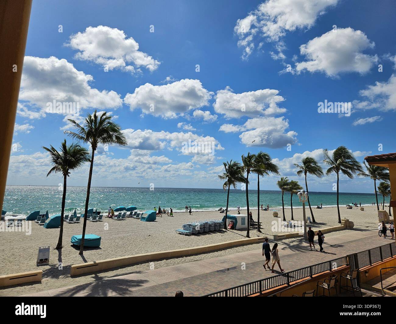 EDITORIAL USE ONLY-HOLLYWOOD BEACH FLORIDA:View from a balcony overlooking Hollywood Beach Broadwalk in Florida. Tourists and locals walking along the oceanfront promenade lined with palm trees, blue beach cabanas, and the Atlantic Ocean under a bright blue sky with scattered clouds. - Smartphone Captured Stock Image
