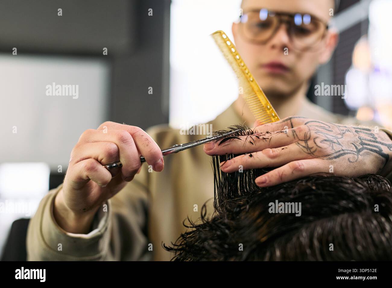 Caucasian young adult man with tattooed hand cutting hair of client using scissors and comb in barbershop, focusing on precise technique and professional grooming service Stock Photo