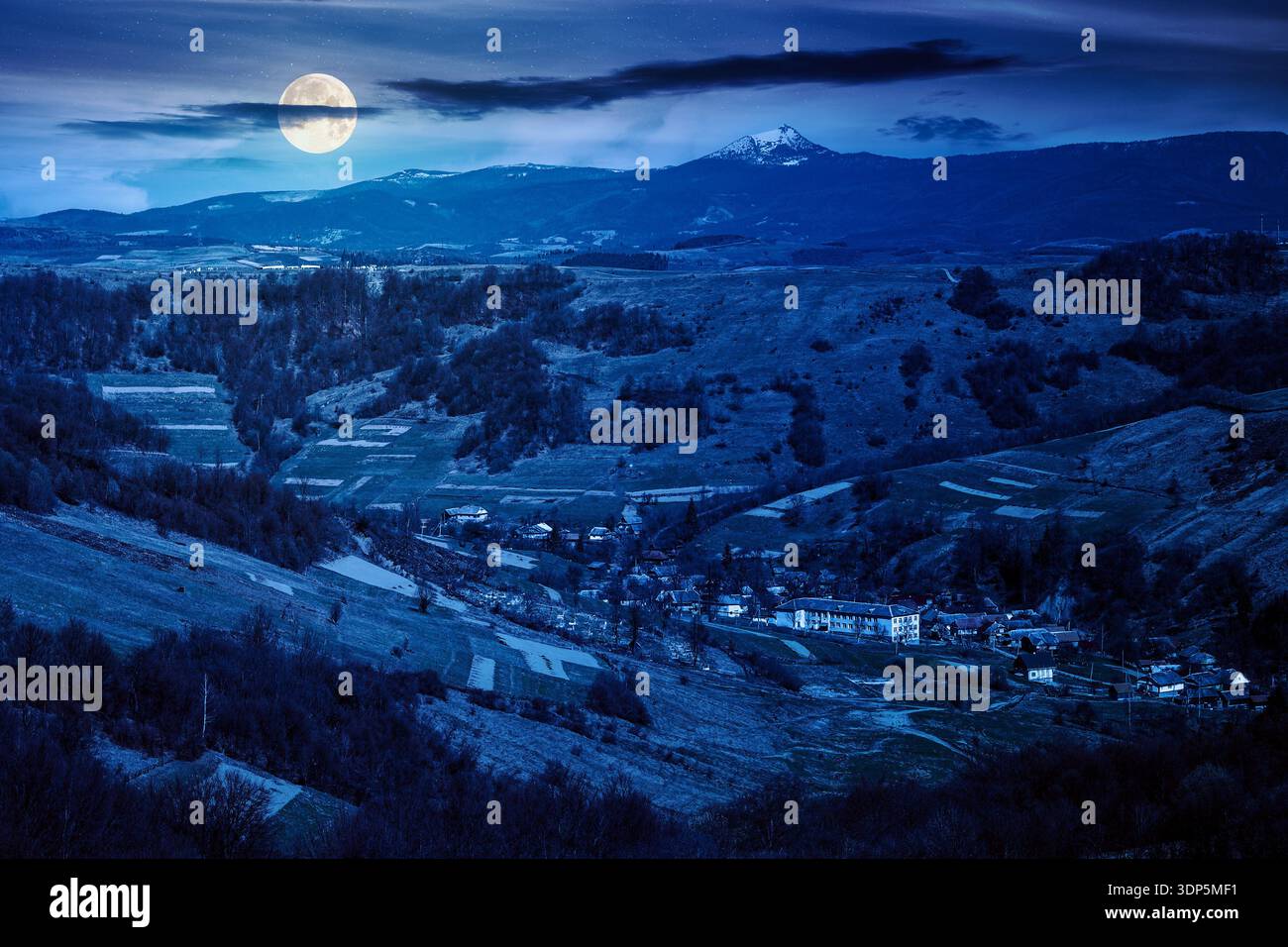 rural landscape with village in valley at night. outdoor adventures in countryside with rolling hills and snowy mountain in early spring in full moon Stock Photo