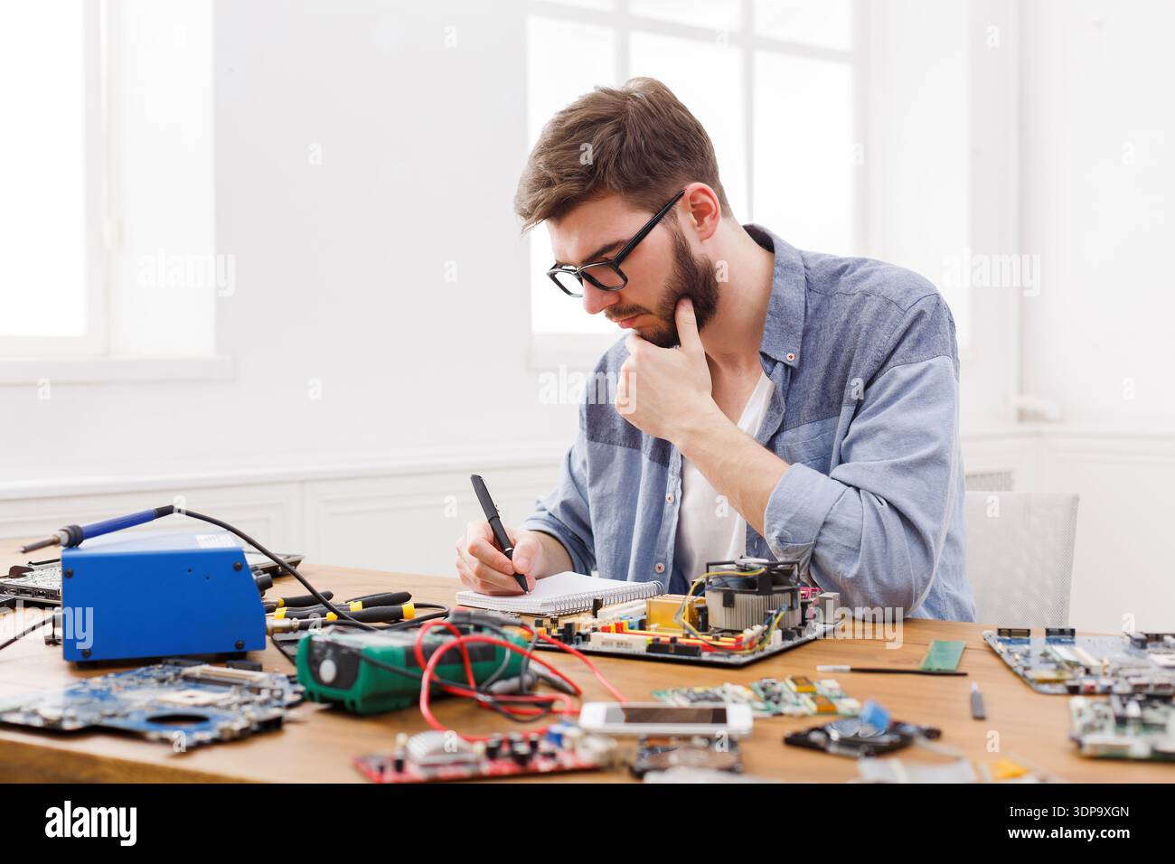 Man Works on Electronics Repair at a Table While Taking Notes in a Bright Room Stock Photo