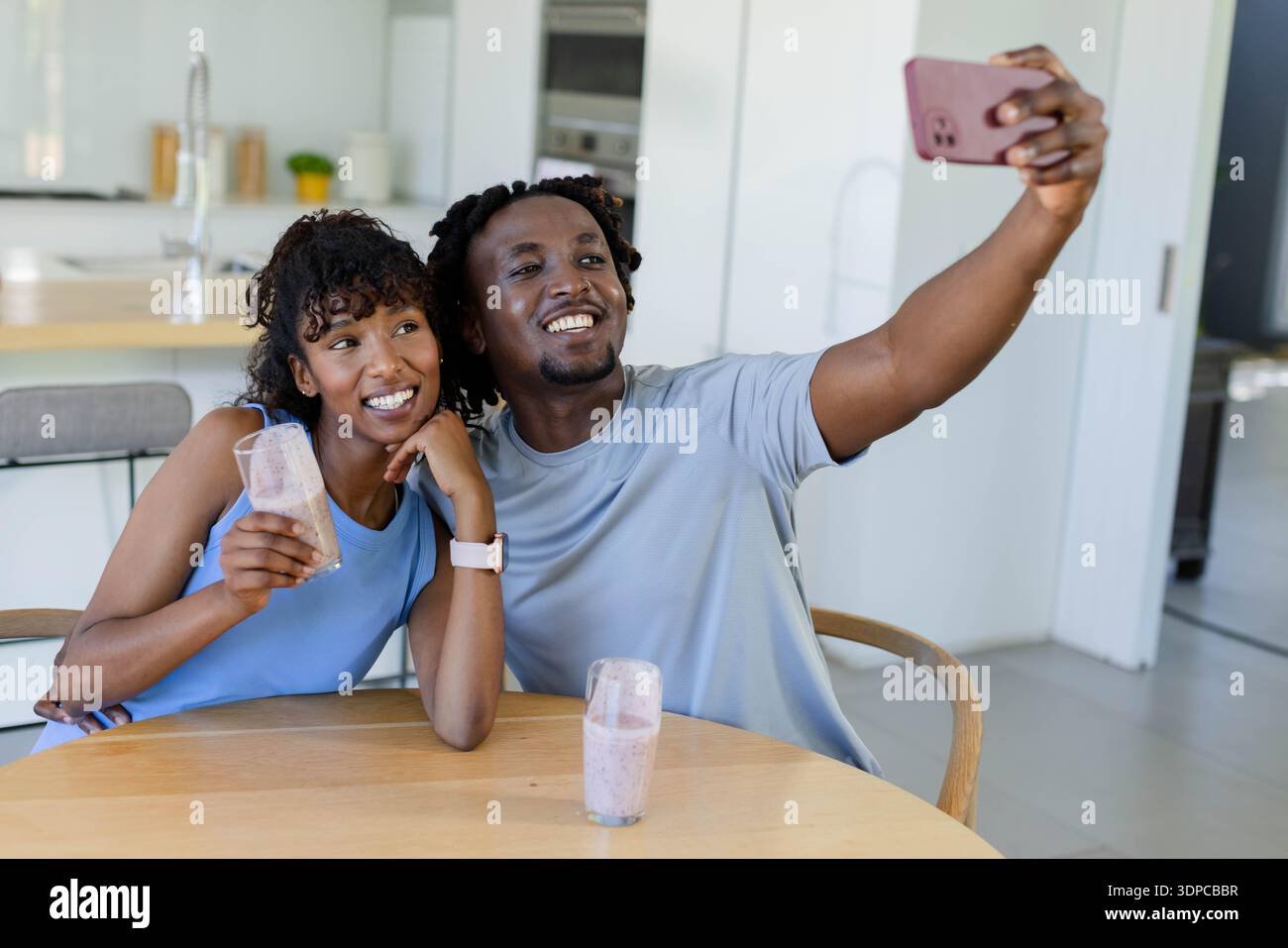 African American couple taking selfie at round table in kitchen, holding pink smoothies, smartphone Stock Photo