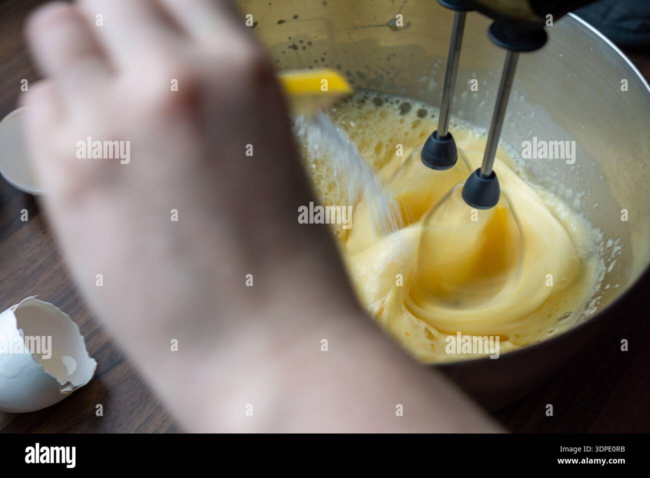 Close up of sugar being poured into egg batter while mixing with an electric mixer in a metal bowl. Stock Photo