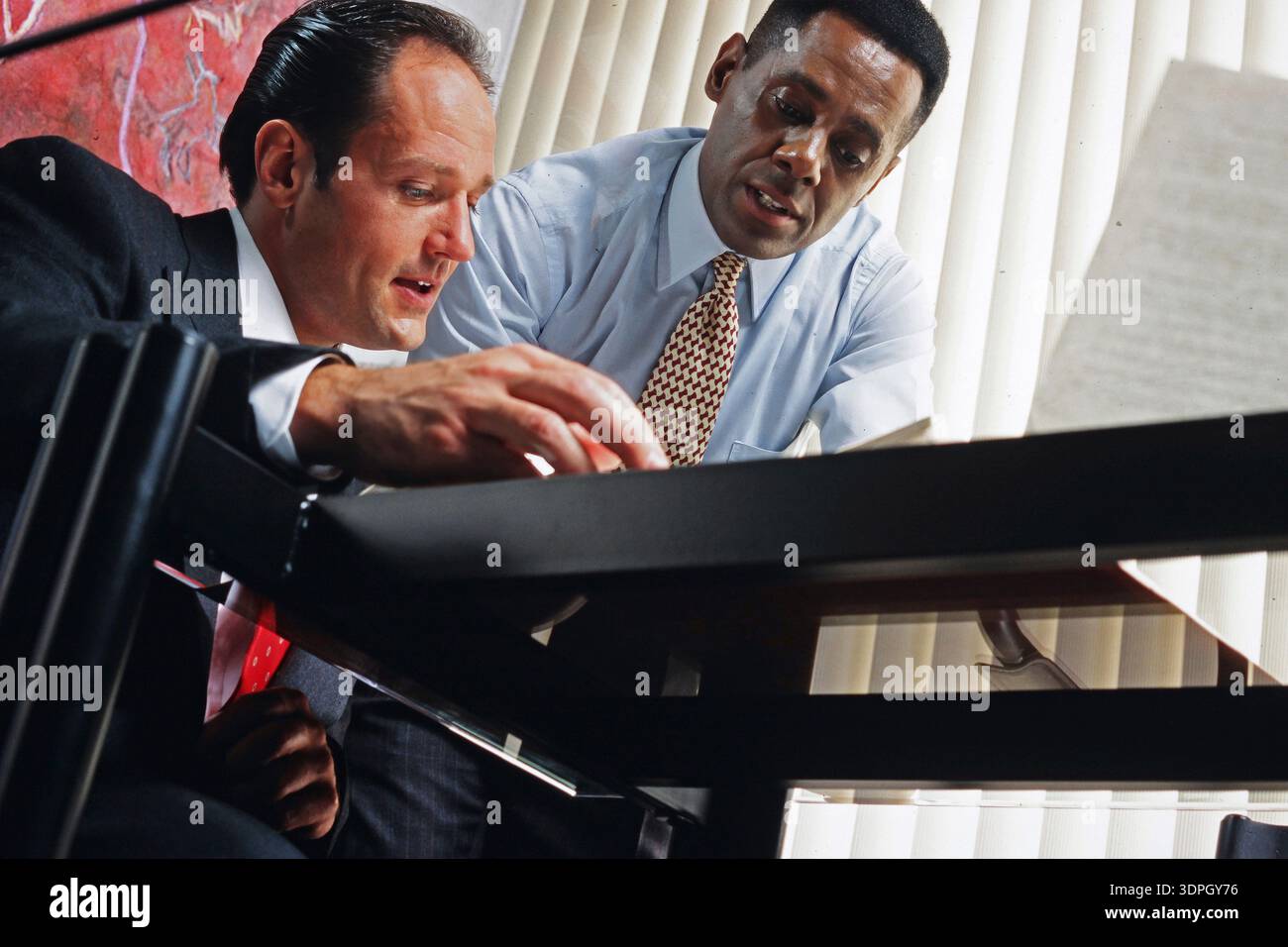 Two men are seated at a table in an office. They are focused on documents and engaged in conversation. They appear to be working on a project together Stock Photo