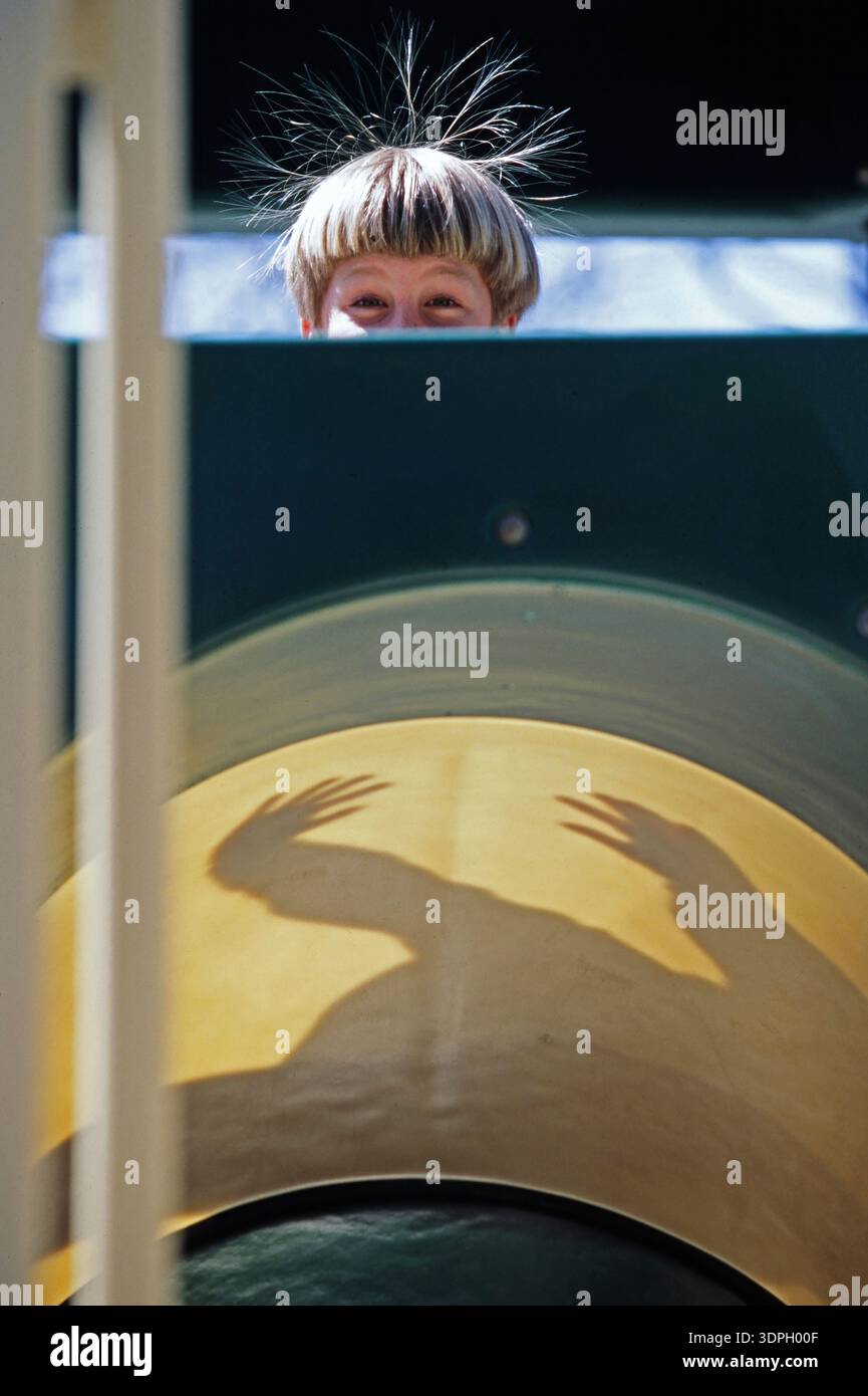 A child with tousled hair peers over the edge of a playground slide. The sunlight casts shadows of playful hand movements. The scene shows joy and ima Stock Photo