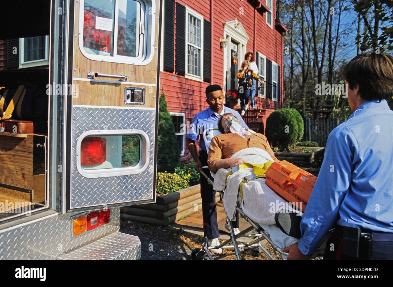 An emergency medical team carries a patient on a stretcher from a house to an ambulance. Family members stand on the porch, observing the situation in Stock Photo