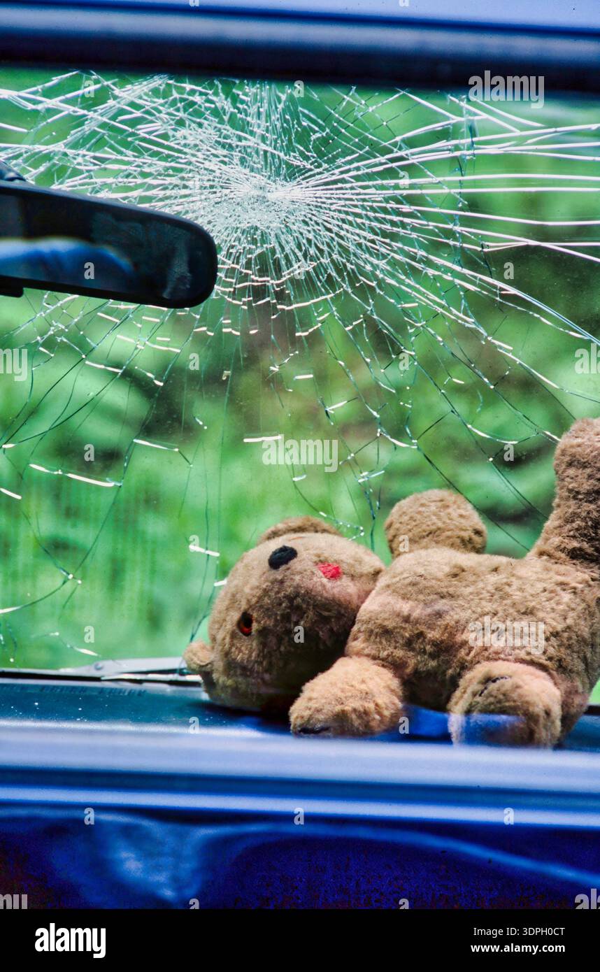 A child's teddy bear lies on the front seat of a blue car. The windshield is cracked and broken after a crash Stock Photo