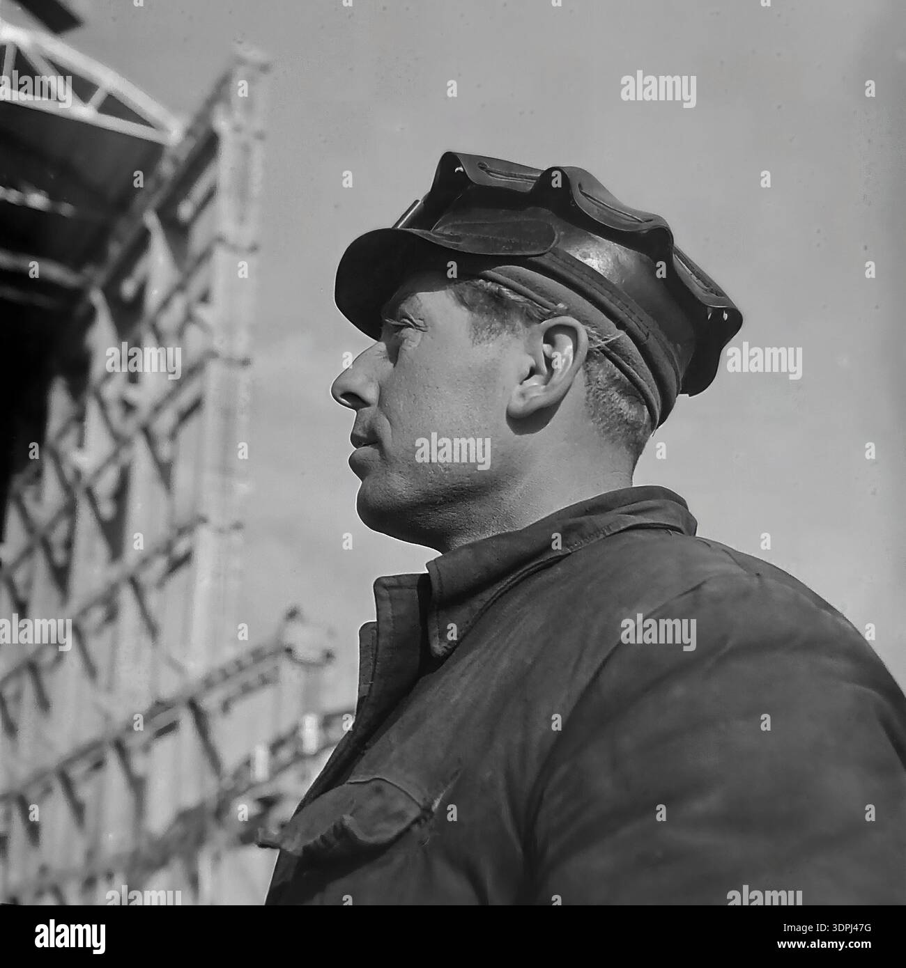 A dramatic archival profile portrait from the 1960s showing a Soviet welder or steelworker at the construction site of the Slavyanskaya Thermal Power Plant (GRES) in Nikolaevka, Donetsk region. The man wears a cap with protective goggles resting on the brim and a sturdy work jacket. He looks resolutely into the distance against a backdrop of a massive steel building skeleton. This authentic close-up captures the strength of the working class and the atmosphere of a peaceful Donbas during the industrial boom. Stock Photo
