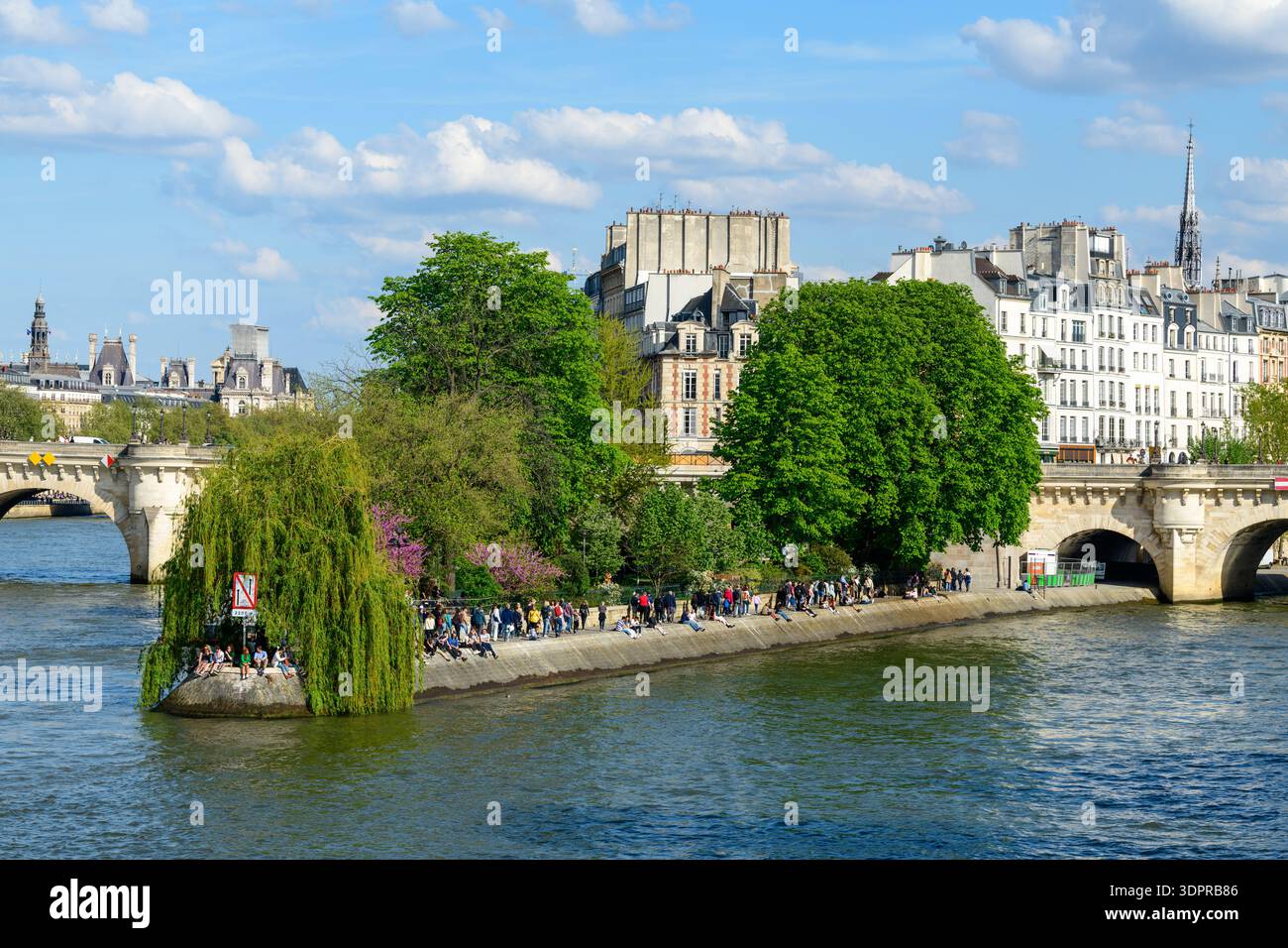 Paris, France - April 14, 2022: Le Square du Vert Galant is shown at the tip of Ile de la Cite with people sitting and walking along the riverbank, lush green trees, Le Pont Neuf bridge, and historic Parisian buildings in the background under a partly cloudy sky. Stock Photo