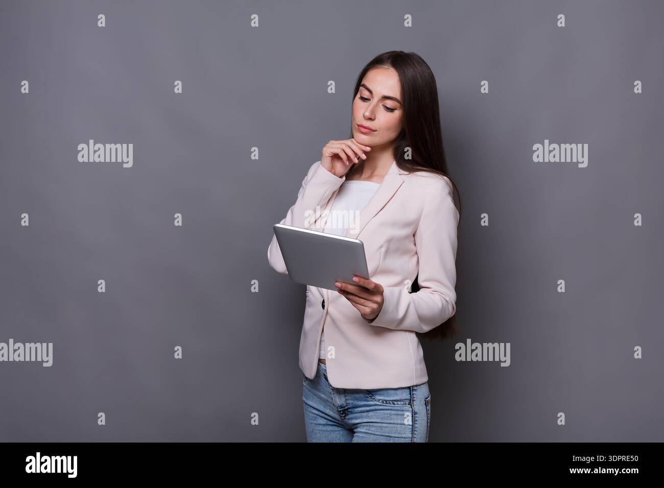 Young Woman in a Light Jacket Thinking While Using a Tablet Against a Gray Background Stock Photo