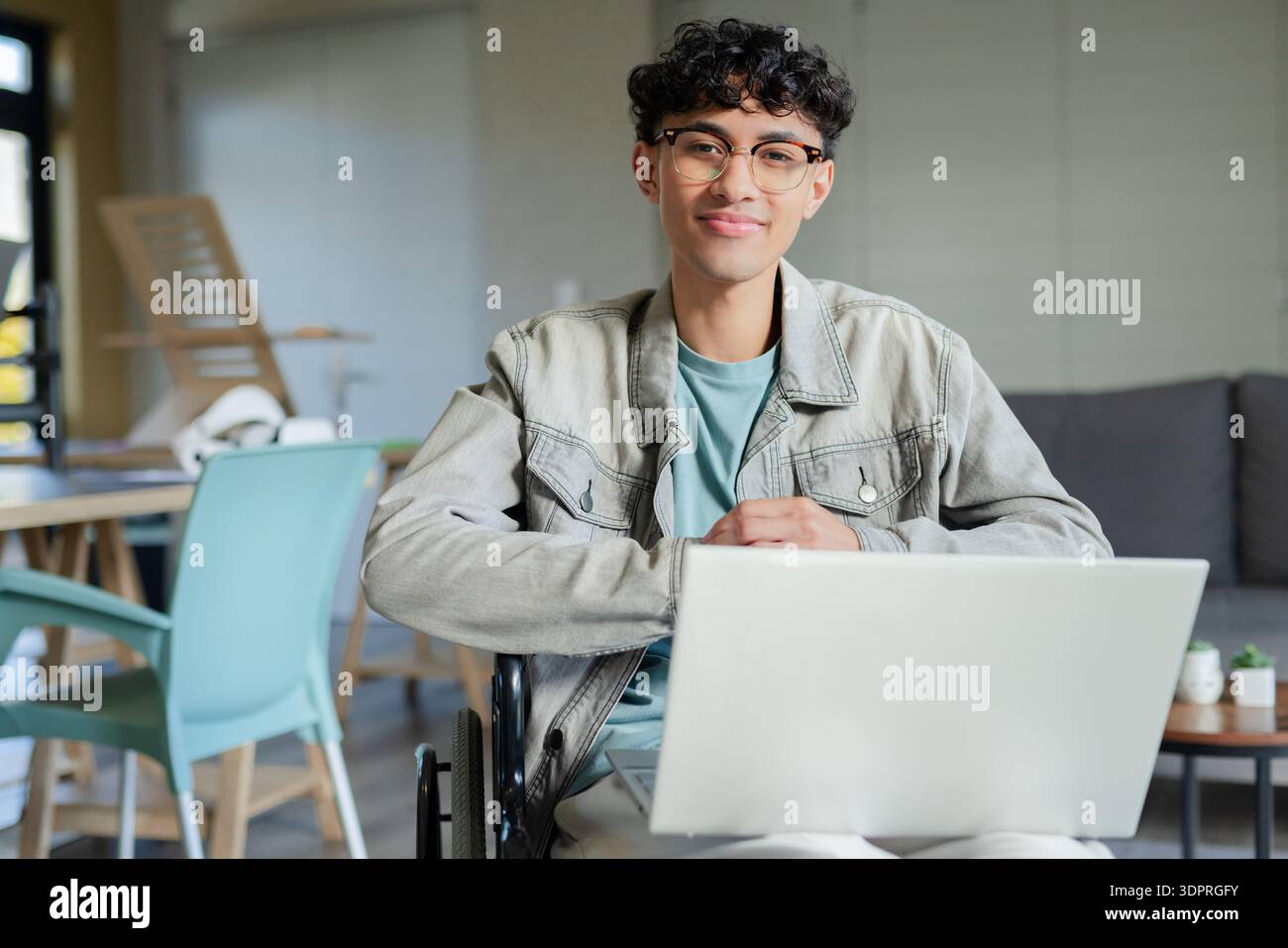 Man sitting in wheelchair using laptop in modern lounge wearing denim jacket and eyeglasses Stock Photo