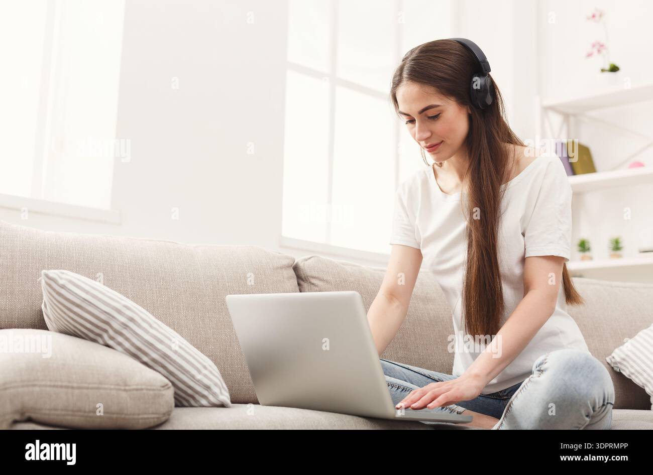 Young Woman Sits on Couch Using Laptop With Headphones in Bright Room During Day Stock Photo