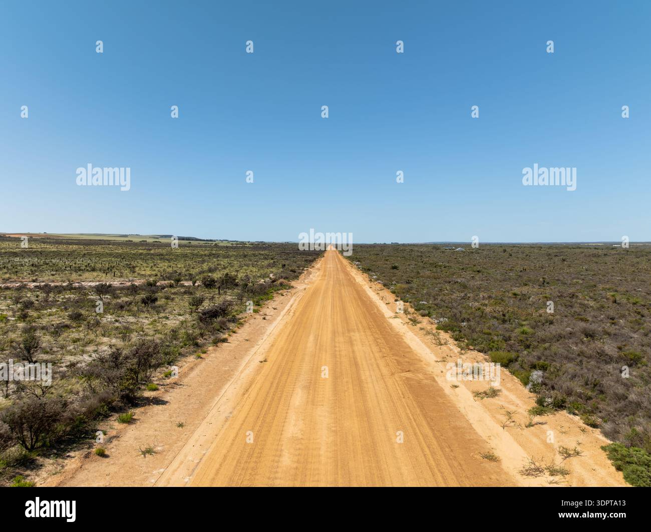 An elevated aerial view of a straight unsealed road stretching through open scrubland and low vegetation under clear blue skies. Stock Photo