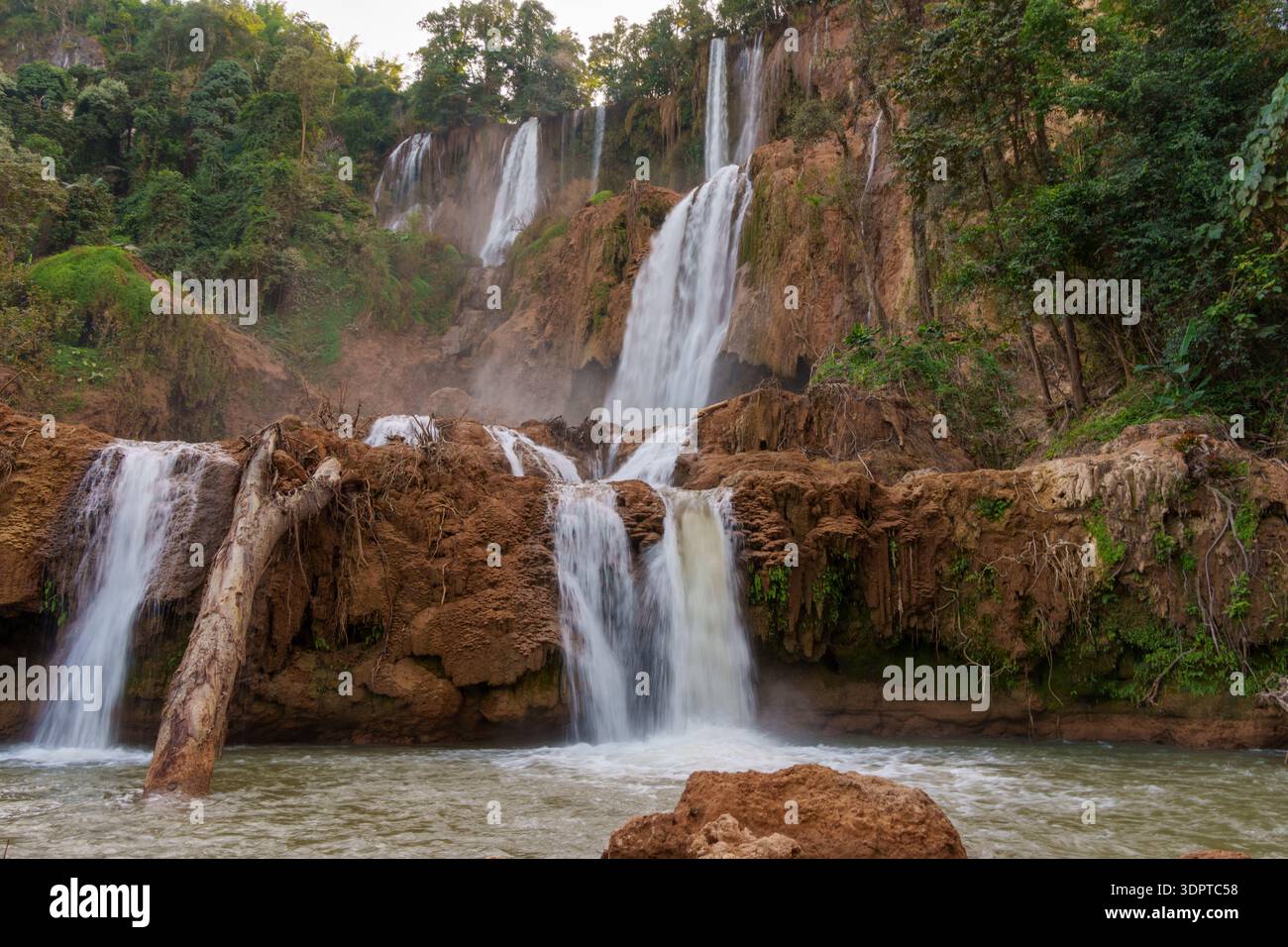 A large multi tier waterfall cascades over brown rock ledges into a fast-moving river, surrounded by dense tropical forest. Stock Photo