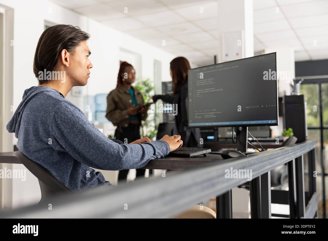 Focused software programmer works in diverse office, ensuring smooth web development. Young adult man types at computer, fully concentrated and committed to completing coding projects efficiently. Stock Photo