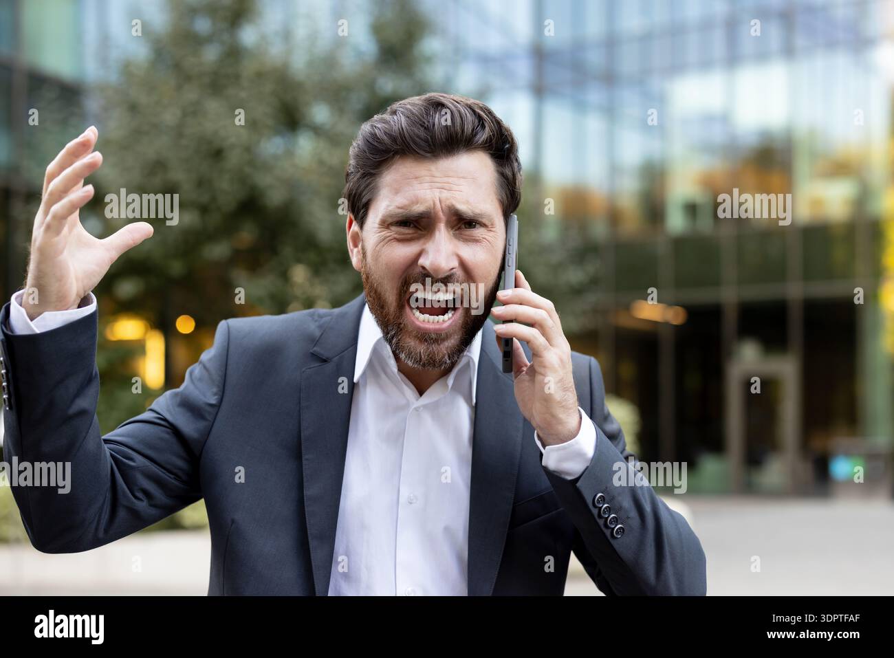 Stressed businessman in a suit having an angry phone conversation outdoors, expressing frustration and anger while using a smartphone in an urban corporate environment Stock Photo