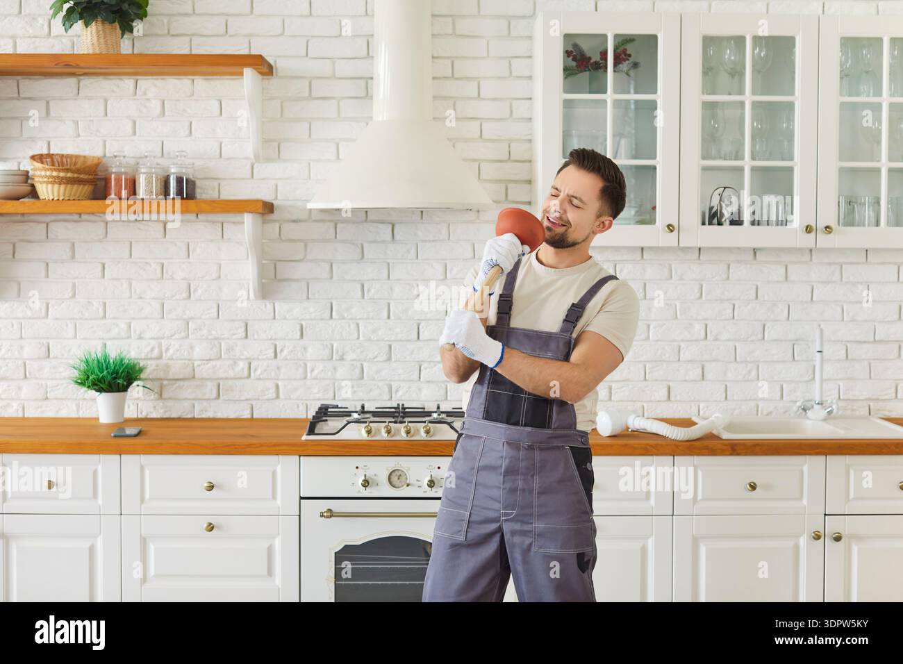Plumber holding plunger like microphone and singing in modern kitchen Stock Photo