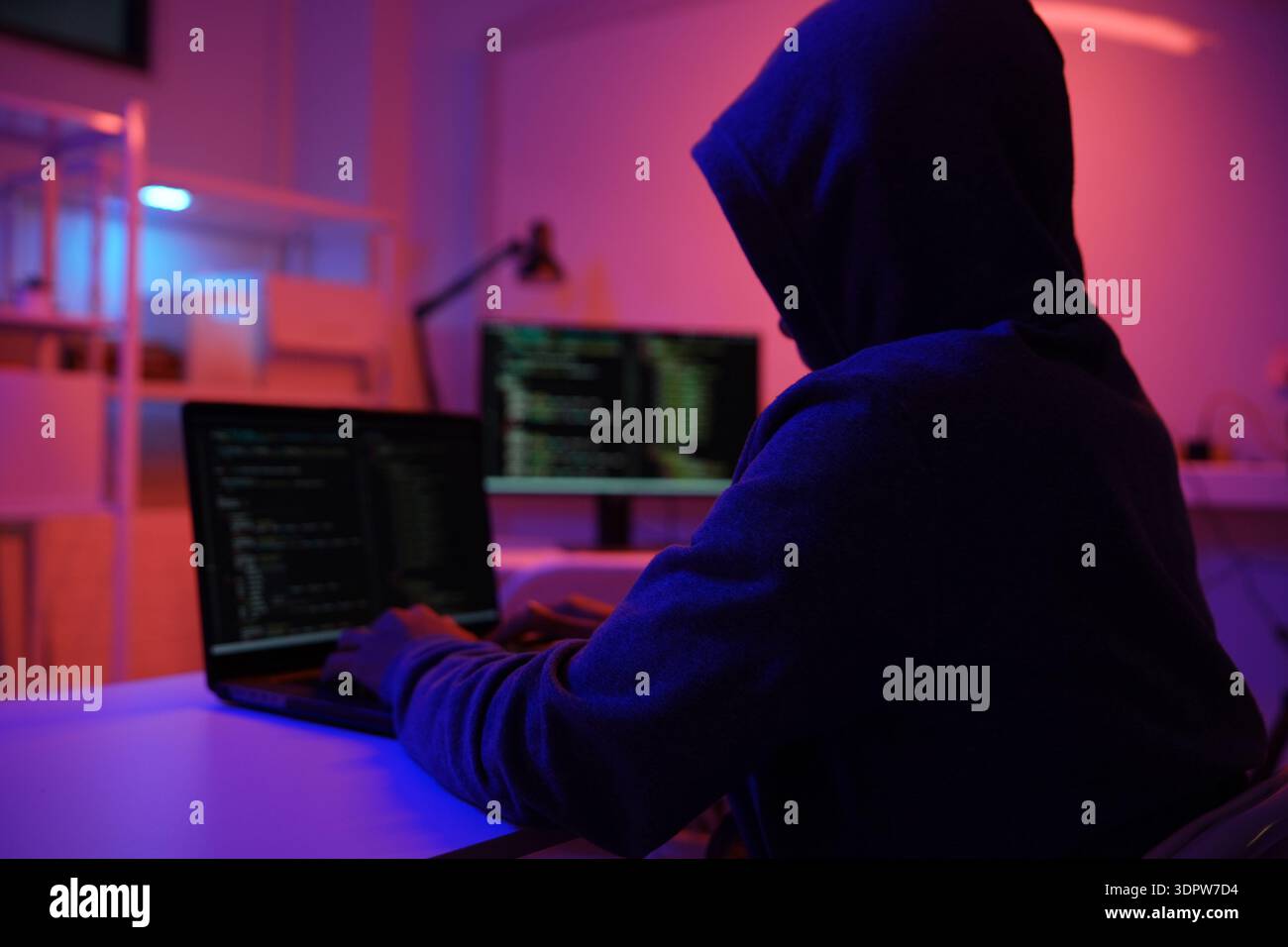A mysterious hooded hacker working on a computer in a dark room. Stock Photo