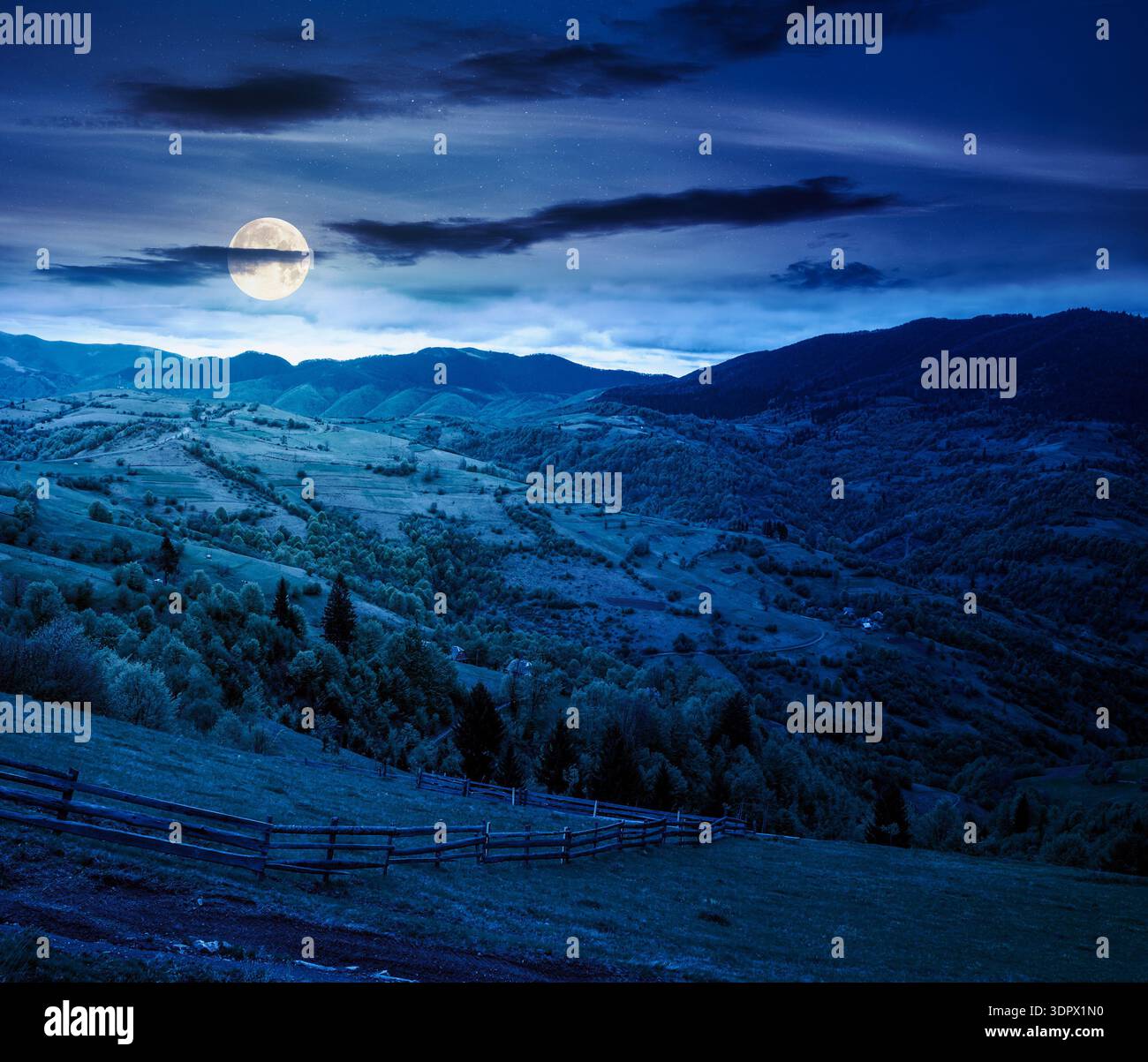 rolling hills of rural landscape in green grass at night. wooden fence in mountains in full moon light. alpine countryside in spring. cloudy sky. back Stock Photo