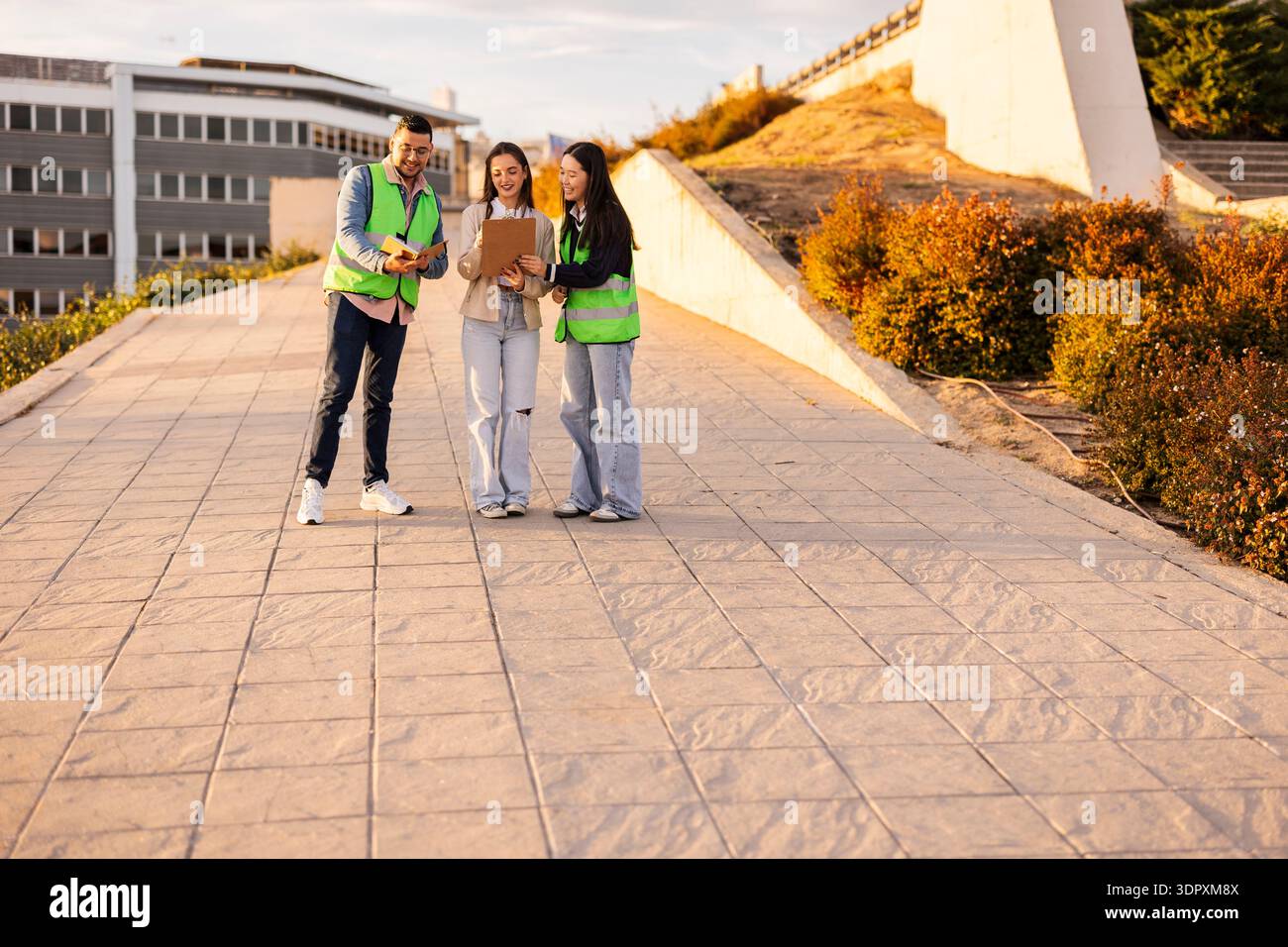 Three students wearing reflective vests discuss their project outdoors, using a clipboard and digital devices under sunny skies on a campus walkway. Stock Photo