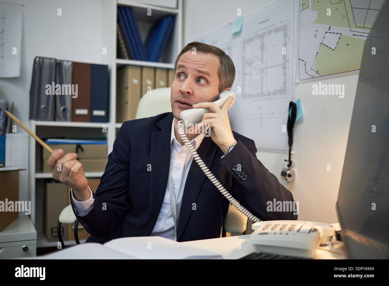 Middle aged Caucasian male engineer or architect sitting at office desk talking on landline phone, holding pencil, gesturing with hand, surrounded by architectural blueprints and documents Stock Photo