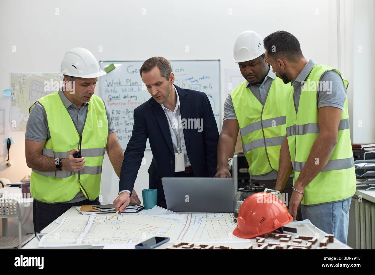 Middle aged Caucasian man in suit collaborating with two middle aged men and one young adult man in safety vests and helmets, discussing construction plans in office, using laptop and blueprints Stock Photo