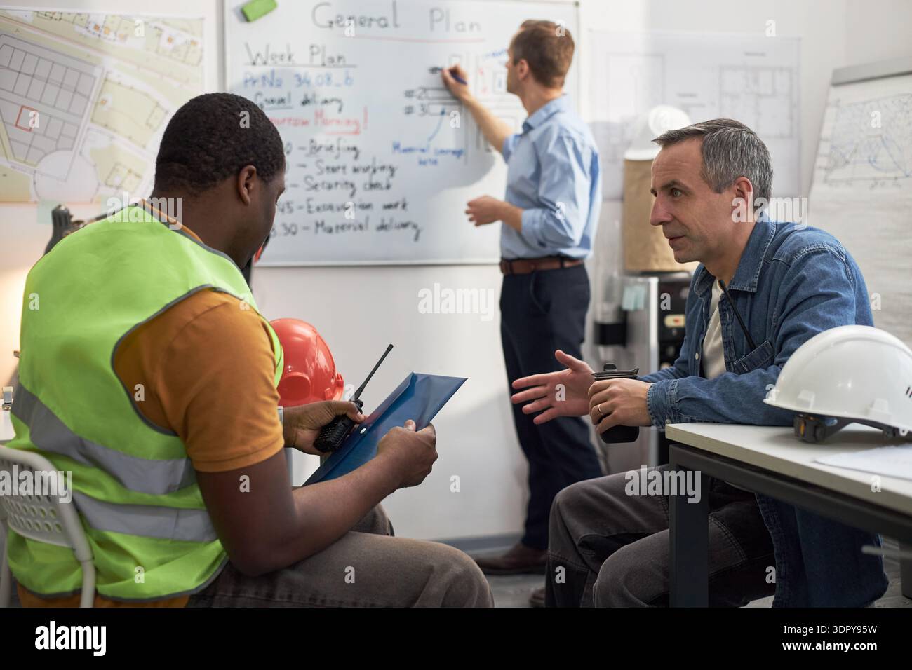 Middle aged Caucasian man discussing construction project details with young adult Black man holding tablet in office, while another middle aged Caucasian man writing on whiteboard in background Stock Photo