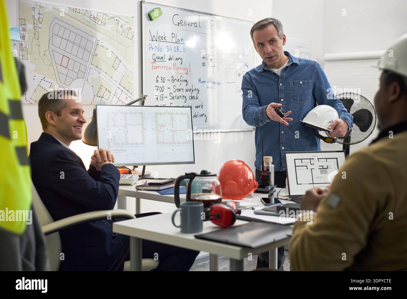 Middle aged Caucasian man standing and gesturing while holding hard hat, discussing construction plans with seated Black man and Caucasian man in office with architectural drawings visible Stock Photo