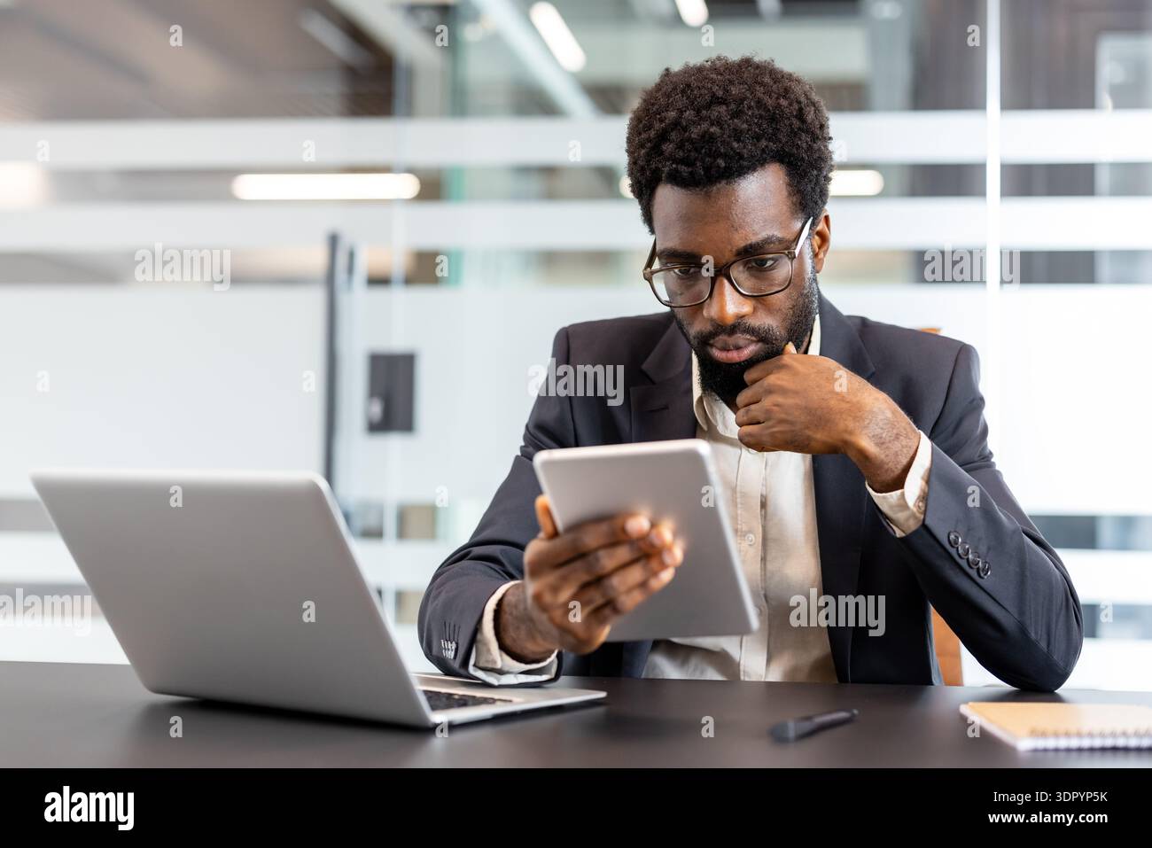 African american businessman sitting at modern office desk, wearing glasses and a formal suit, using a digital tablet while concentrating on a professional task Stock Photo