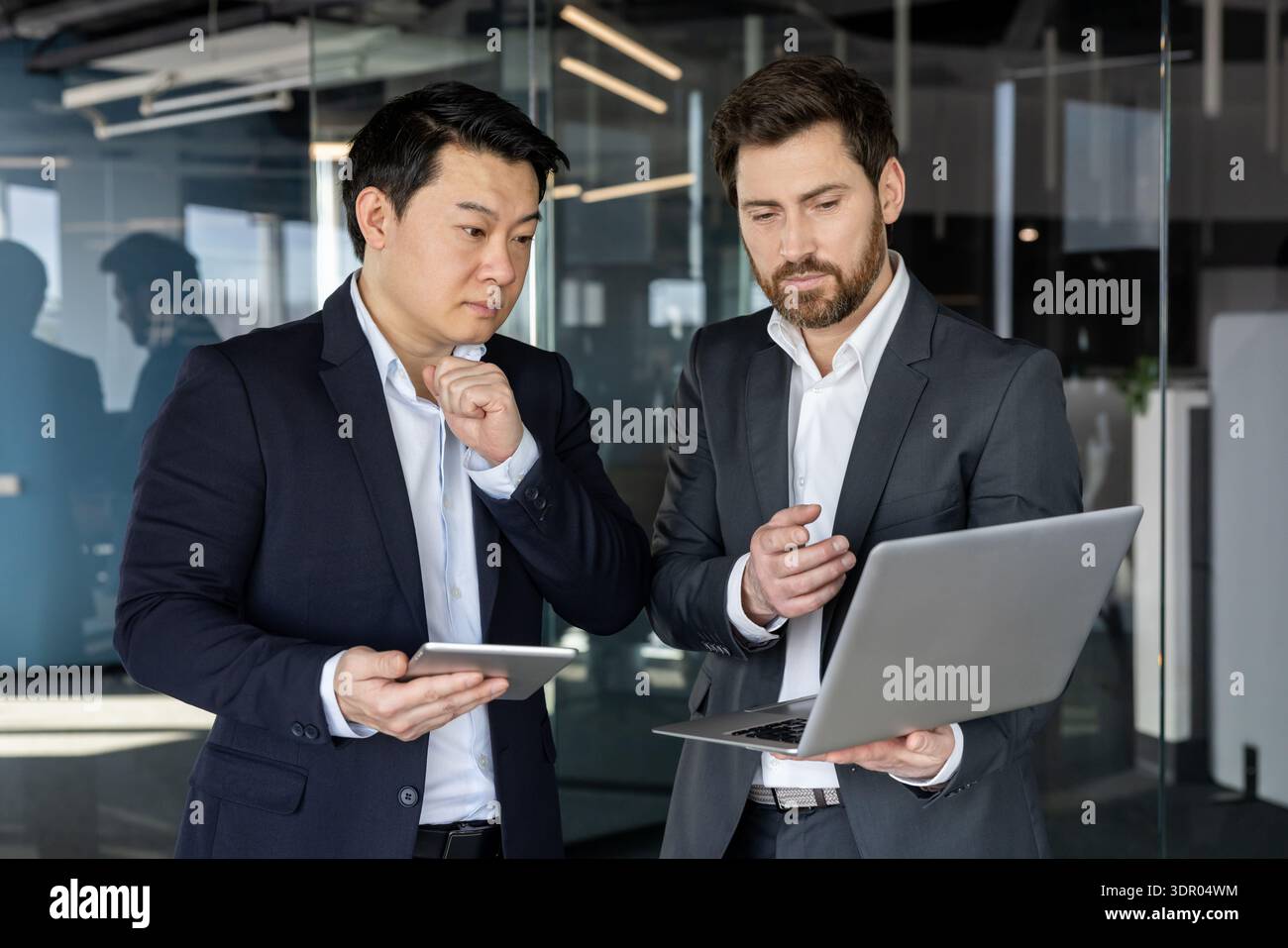 Two diverse businessmen standing in a modern office, one holding a laptop and the other a tablet, collaborating and discussing business strategy while analyzing data Stock Photo