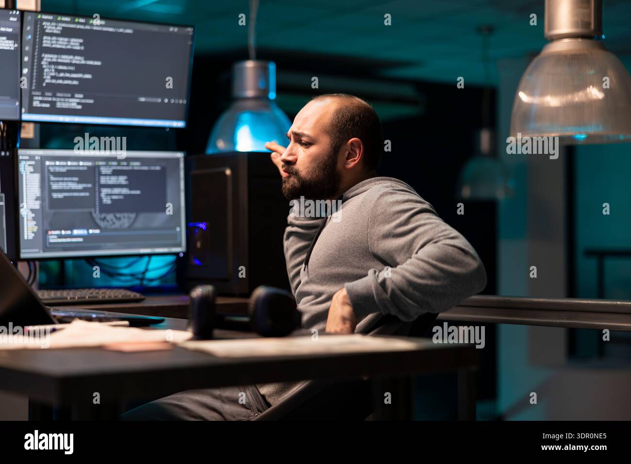 Male coder sitting and dancing with satisfaction after solving complex programming issue. Tech professional enjoys well earned victory in modern office lit by glowing computer monitors. Stock Photo
