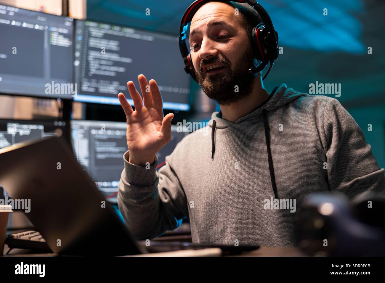 Caucasian IT specialist wearing headset speaks during virtual meeting at desk. Multiple digital screens show coding software as programmer supports clients and collaborates on technical solutions. Stock Photo