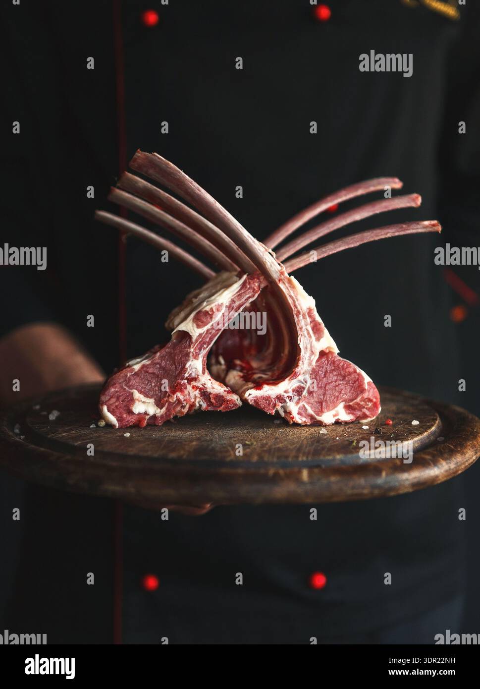 Collection of Raw Lamb Chops Displayed on a Wooden Plate by a Chef in a Dark Kitchen Setting Stock Photo