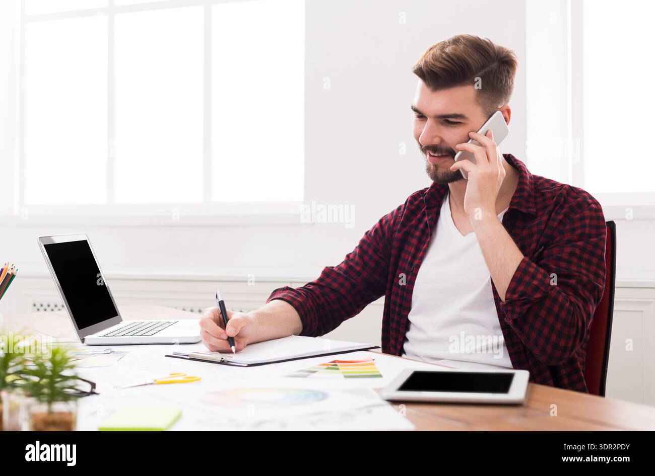 Young Man Working at a Desk While Talking on the Phone and Taking Notes Stock Photo