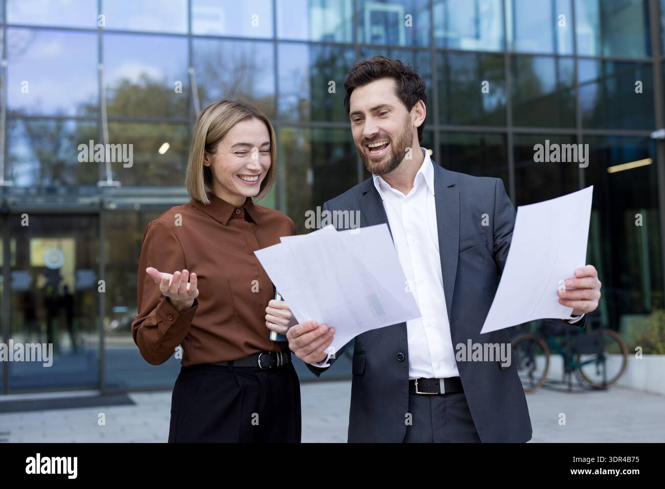 Two smiling business professionals engaging in a lively discussion outside an office building, reviewing paperwork and collaborating on a project with shared enthusiasm Stock Photo