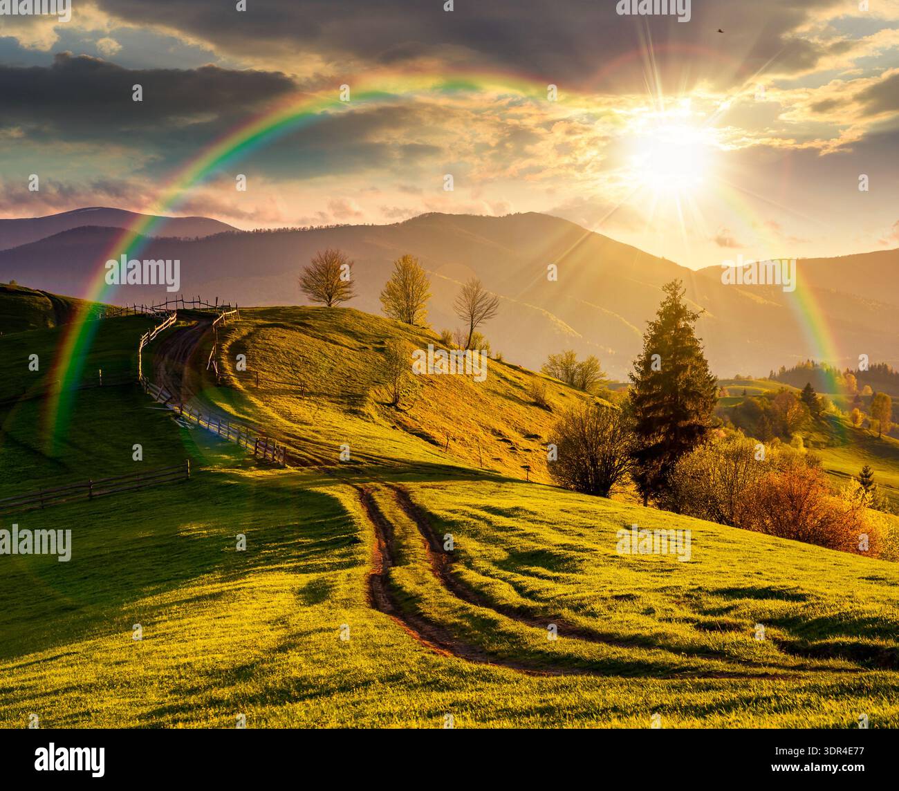 countryside scenery with grassy slopes at sunset. wooden fence along the winding dirt road through rolling hills in evening light. spectacular landsca Stock Photo
