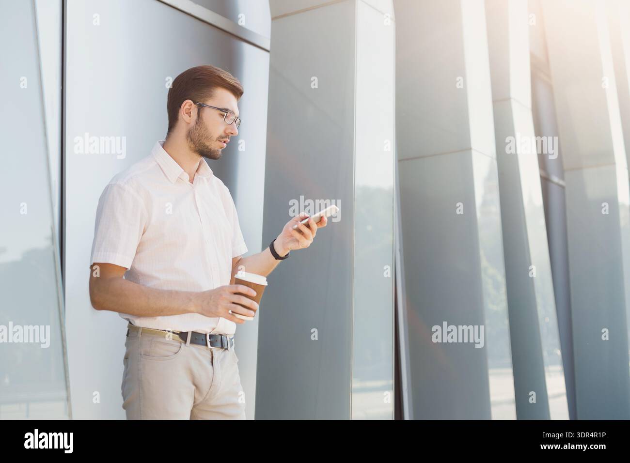 Man Stands by Modern Building While Checking Smartphone and Holding Coffee Cup in Hand Stock Photo
